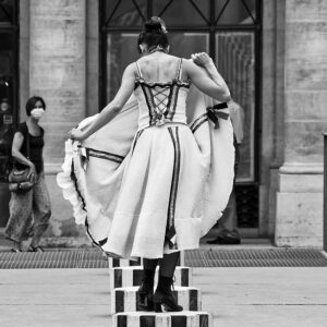 Dancer in black and white seen from behind, lifting her skirt in front of a passerby in the courtyard of the Palais Royal
