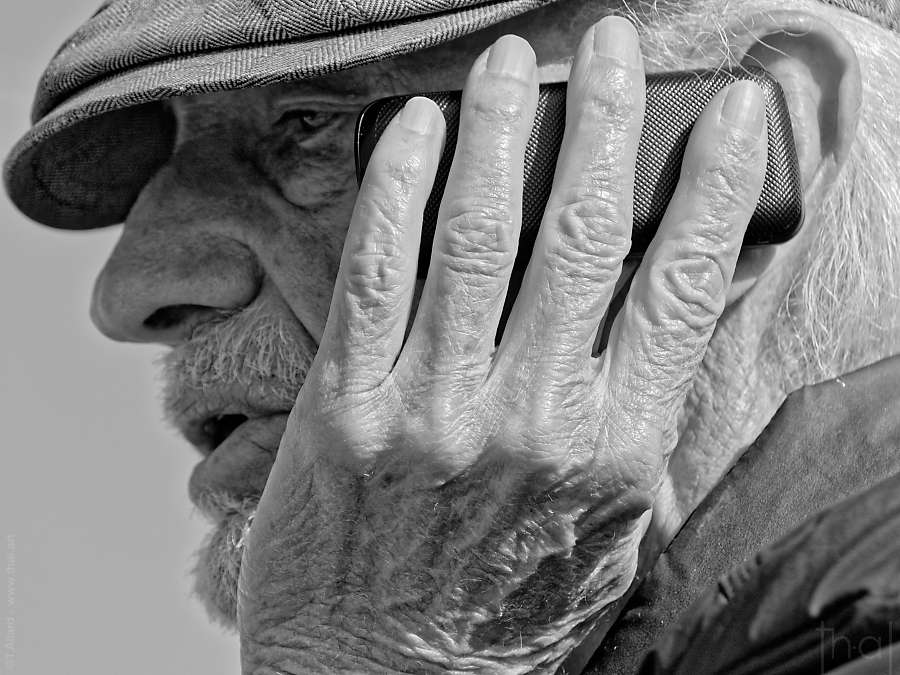 photographie en noir et blanc d'une main ridée plaquant un téléphone sur le visage d'un viel homme